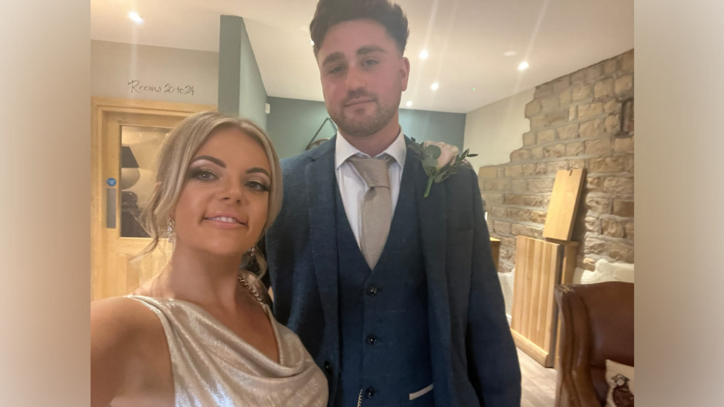 A young woman with blonde hair and a glittery dress, stands next to a young man in a navy suit inside a pub