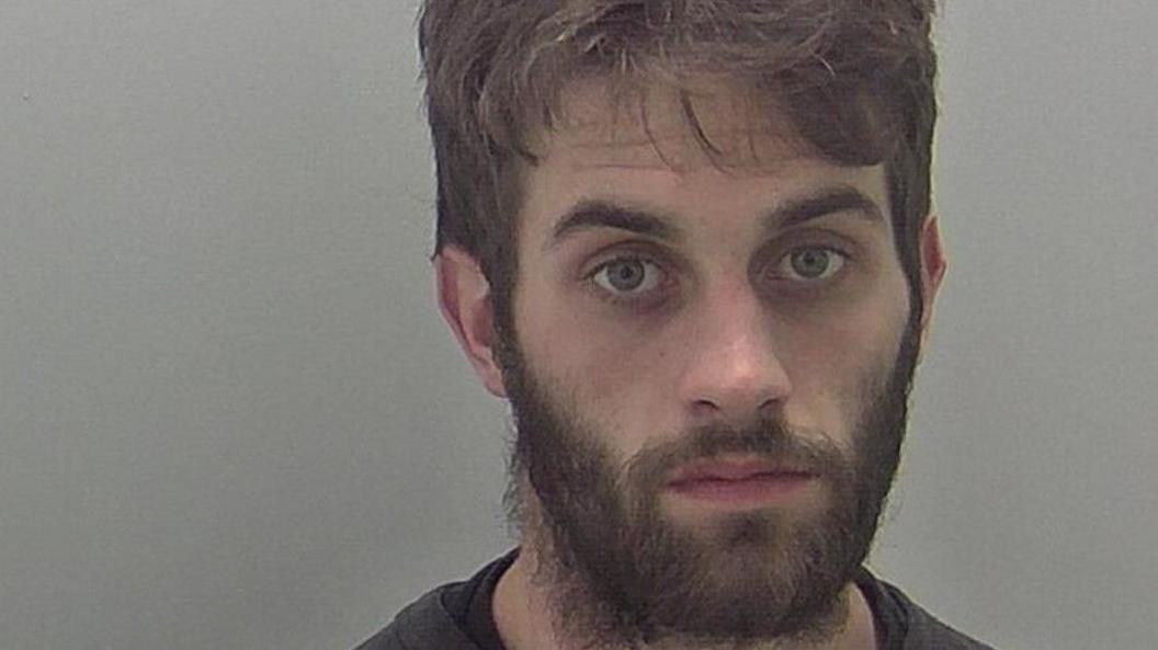 A man with dark hair and a beard is looking at the camera.