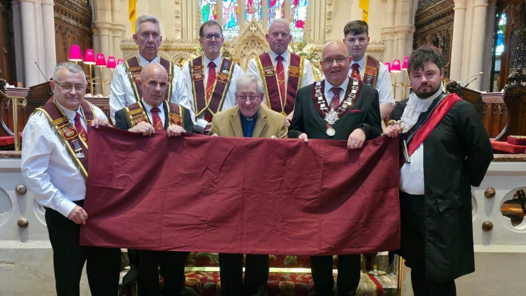 Shows a church minister and members of the Apprentice Boys in full regalia holding a crimson-coloured flag