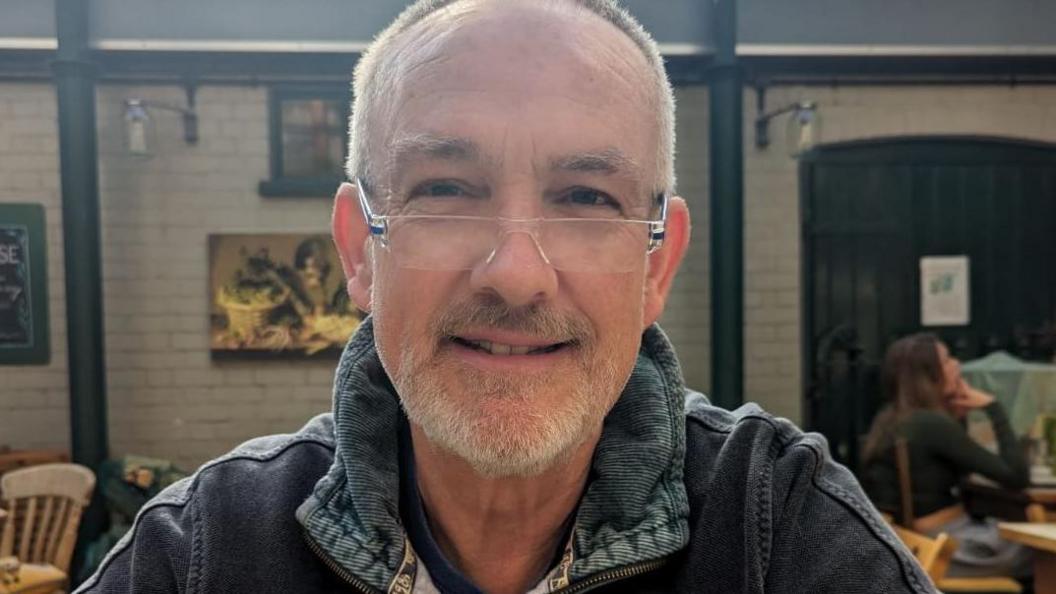 Andrew Sadler smiles at the camera as he is seated in a cafe. He is wearing a denim jumper and glasses. He has white hair and a white beard. 