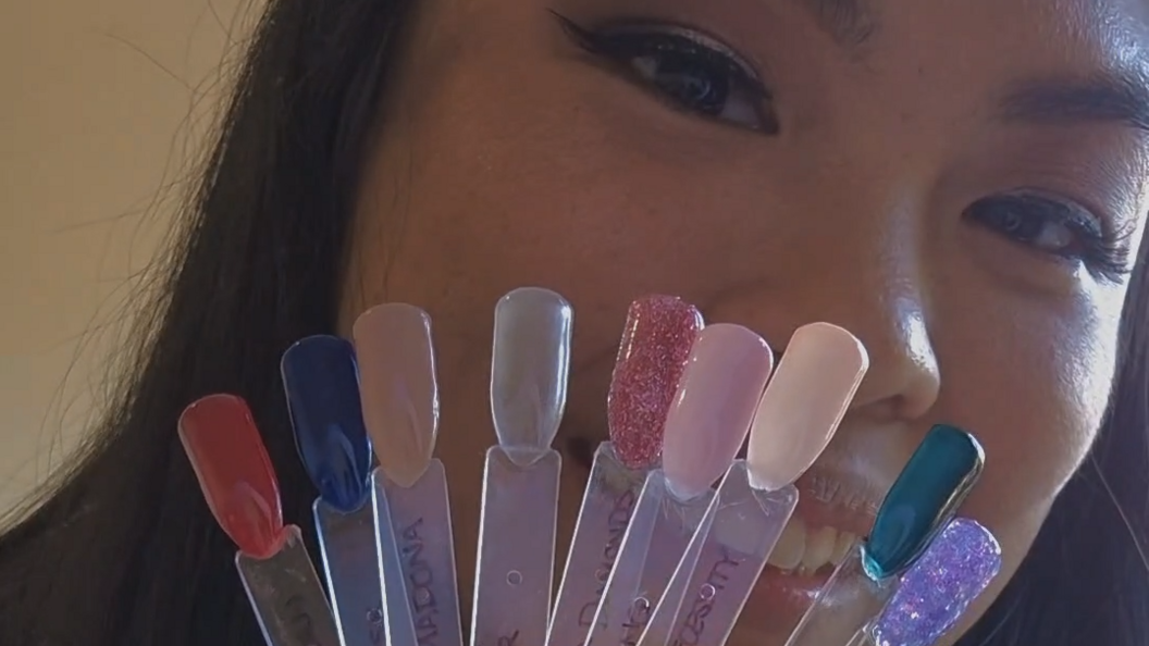A close up shot of a woman smiling. She has black hair and is holding up fake nails on plastic sheets which she has painted in shades ranging from red to blue.