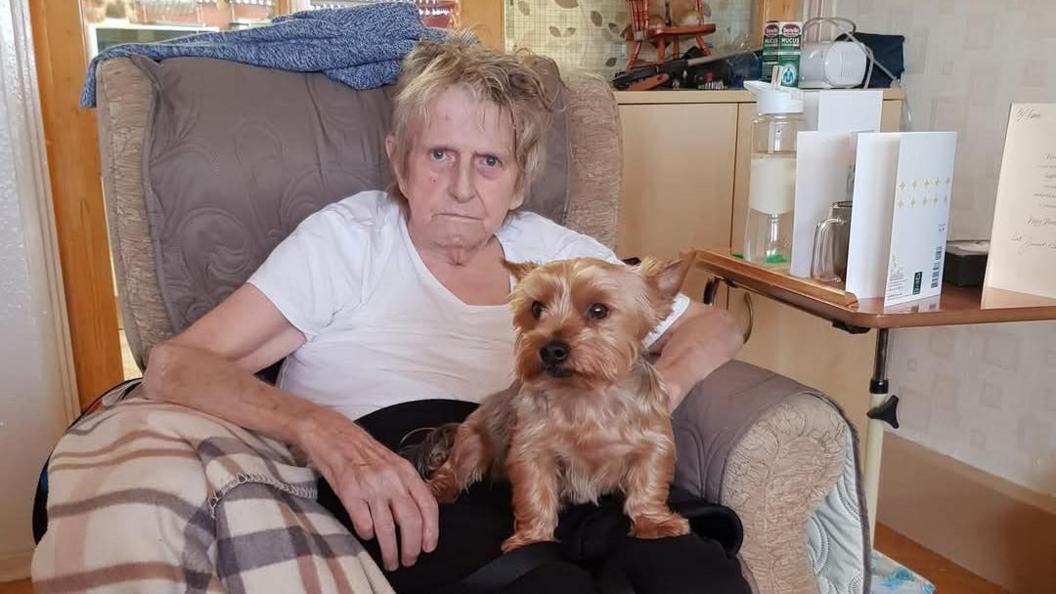 Lois' father, Frank, is sitting on a chair with a little dog on his lap. He is an elderly man who looks unhappy. He is wearing a white T-shirt. There is a table next to him with cards on it.