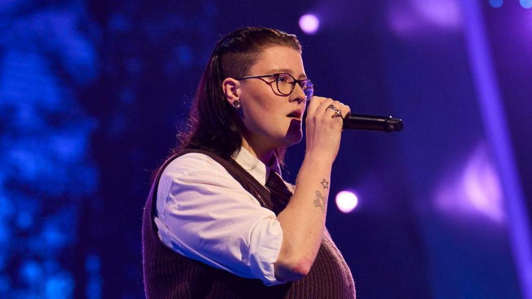 Hollie Peabody performing on The Voice. Hollie has long dark hair and an undercut above their ears. They wear a maroon knitted vest over a white shirt, their sleeves rolled up to their elbow revealing two tattoos: a small star and crossed plasters. They have a serious expression as they hold the mic to their face. 