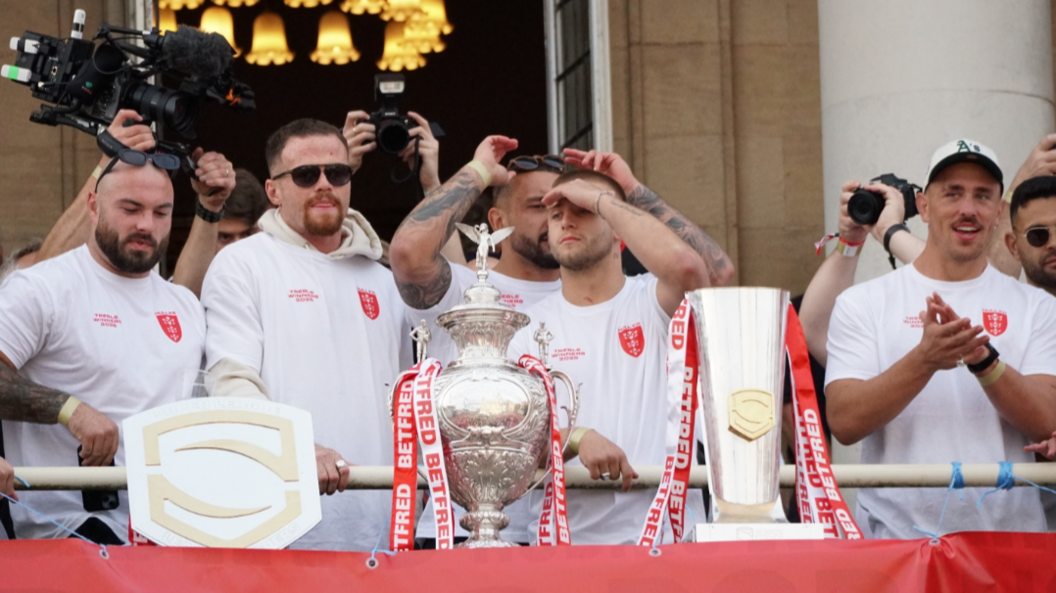 Three trophies are on display on the balcony with read and right ribbons on them.  The players stand behind the trophies on the balcony in white t-shirts with red emblems on them.