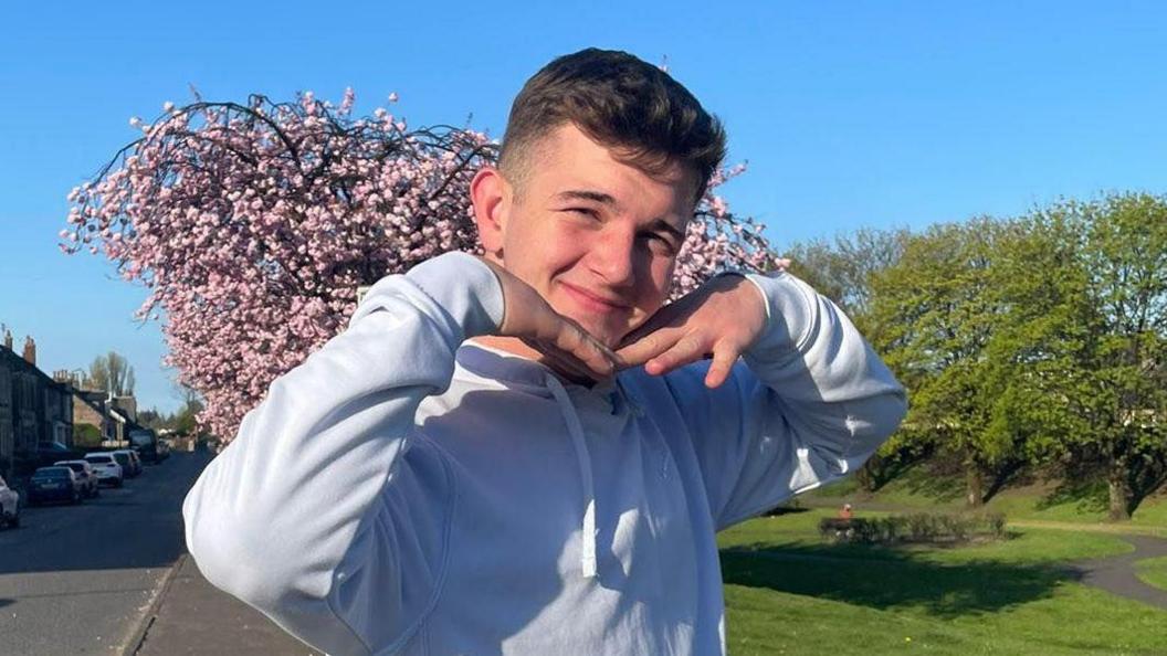 Cole Cooper, a teenage boy, wearing a white hoodie and has his hands placed under his chin in a v-shape as if he is framing his face. He is standing on a pavement next to what looks like a park with grass and a blooming cherry blossom tree behind him.