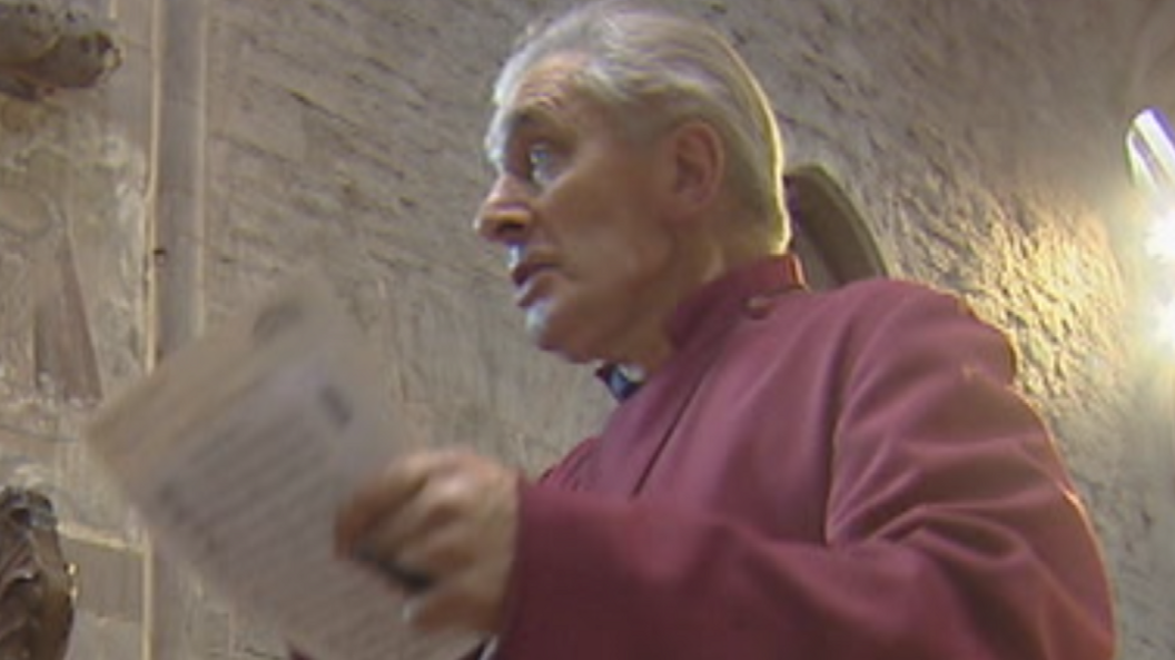 A white-haired man with a purple robe inside a church holding a piece of paper is speaking