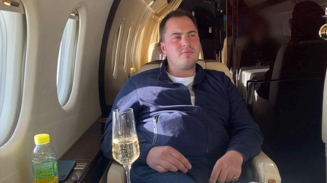 Miles Hart, a young man with short brown hair, wearing a quarter-zip blue top, sitting in a cream leather seat on a small, luxurious jet, with a glass of what appears to be champagne in front of him.