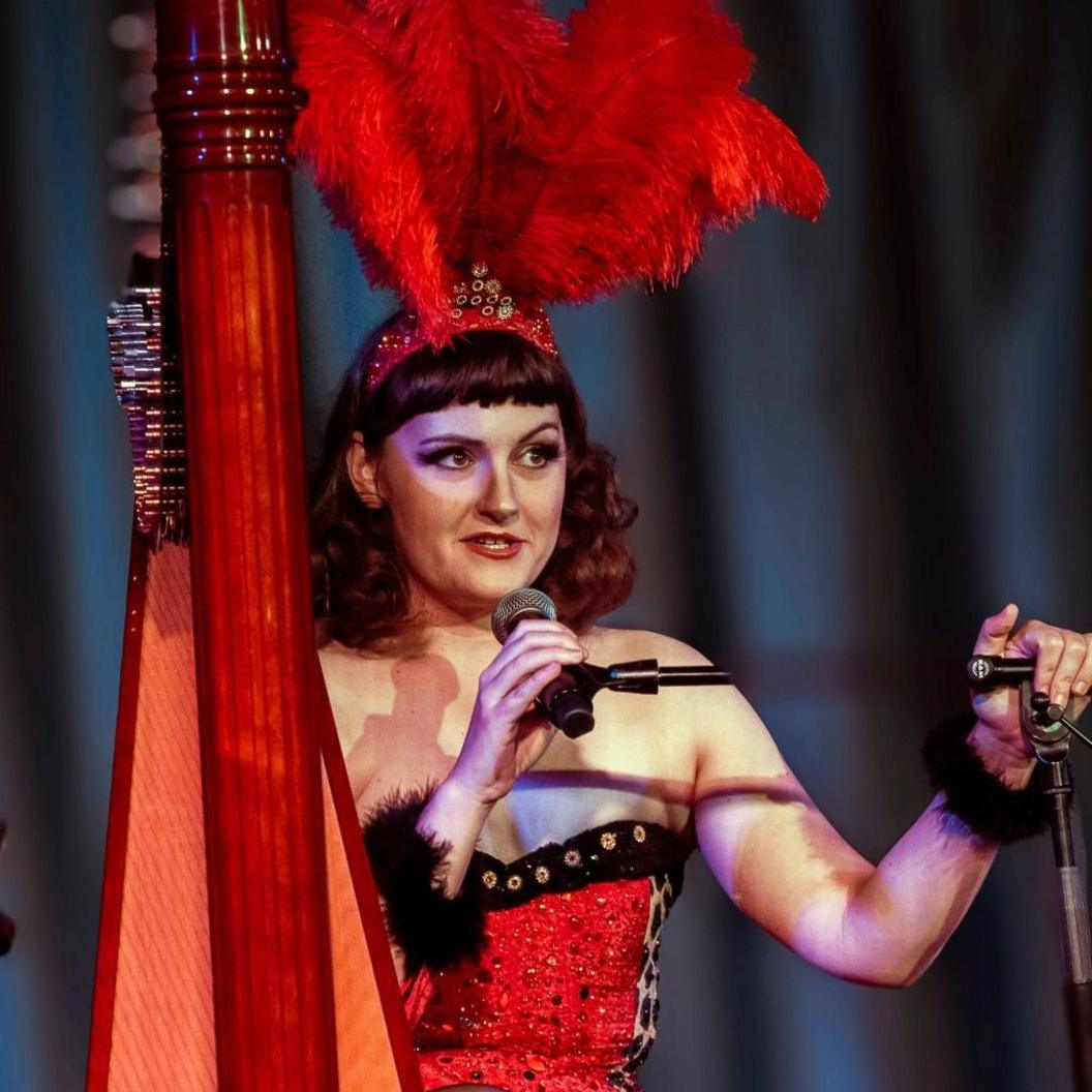 Sam Hickman mewn dillad coch o flaen telyn