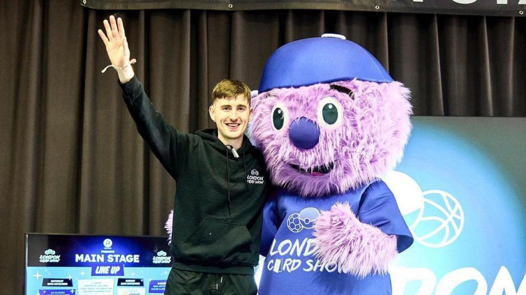 Harry Reynolds, wearing a black hoody and jogging bottoms, stands on stage with another person dressed as a purple bear. Harry has his hand in the air and is waving.