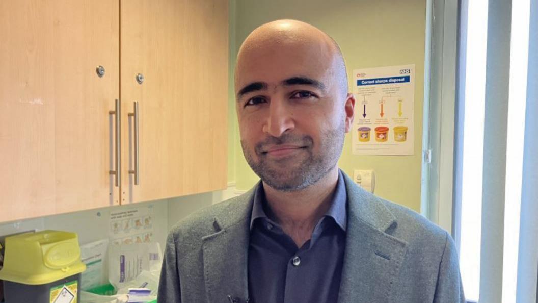 A bald man with a faint beard in a grey suit in a room with wooden cupboards on one wall and a small yellow medical waste bin on a worktop