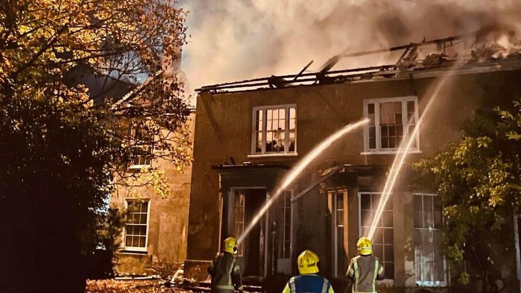 A georgian-style house with smoke billowing out the top - roof destroyed, walls standing but appears  mostly destroyed inside too. Firefighters outside with hoses.