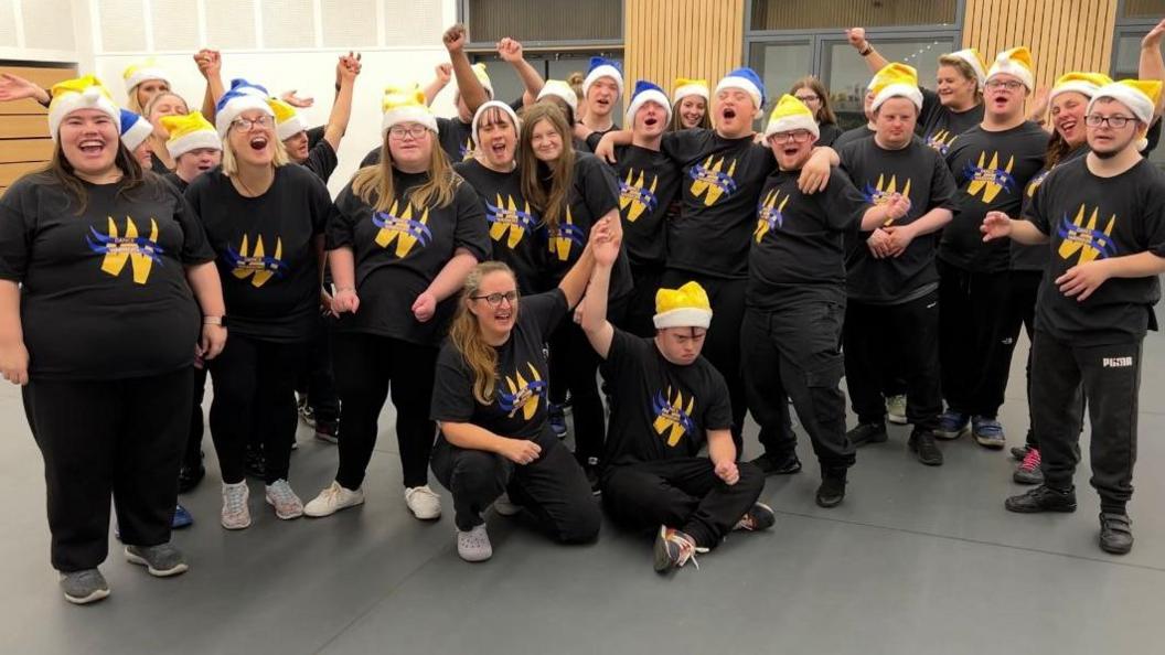 A group of dancers with learning disabilities are stood having their photo taken together. Some are cheering and smiling. 