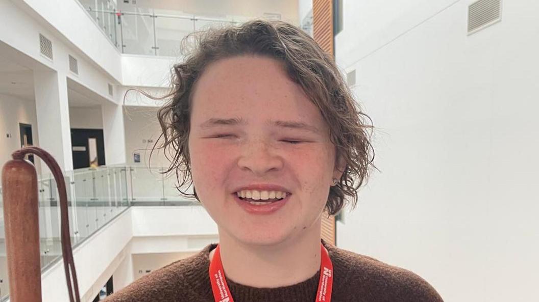 A girl with long brown hair wearing a brown jumper and a red University of Northampton lanyard stands in a stairwell. 