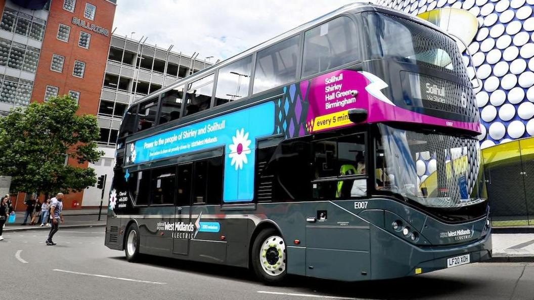 A National Express bus near the Bull Ring in Birmingham, with the shopping centre's silver, shiny circles visible on the outside. There are pedestrians walking around and there is car park behind the bus.