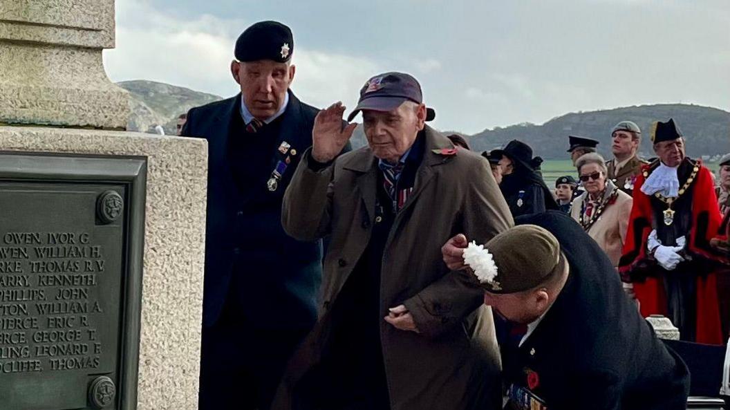 Richard Jons pictured standing in front of a remembrance statue. He holds his left hand in the air to salute, a solider holds his right arm. He wears a dark green trench coat, and another soldier stands to his left.