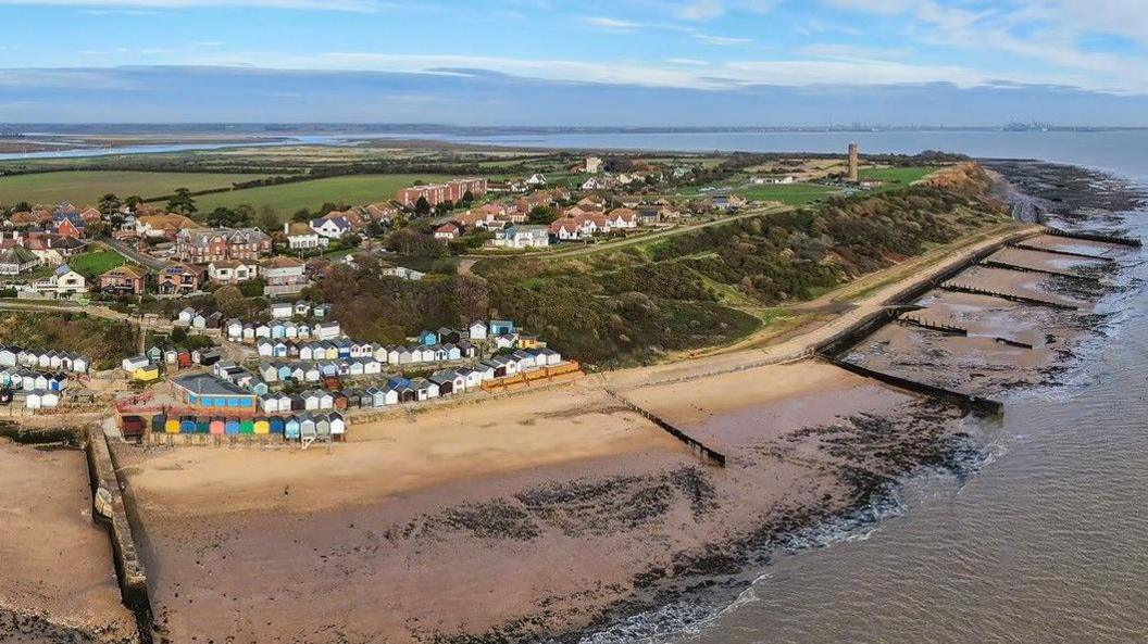 An aerial picture taken of part of the beach at Walton-on-the-Naze. You can see beach huts and houses, as well as the sand and sea. In the background, you can see the Naze Tower atop of the clifftop