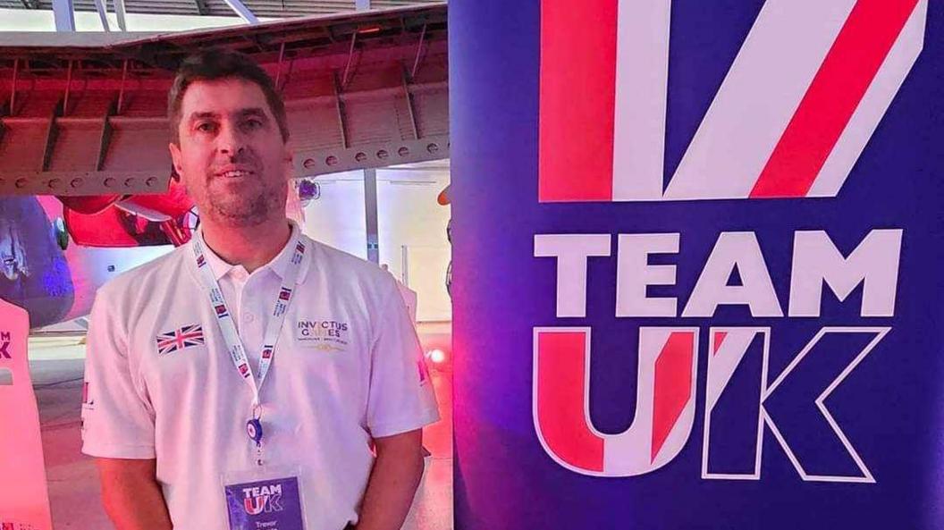 Mr Bygate wearing a white polo shirt with "Invictus Games" logo and a lanyard around his neck, stands beside a poster that reads "Team UK"