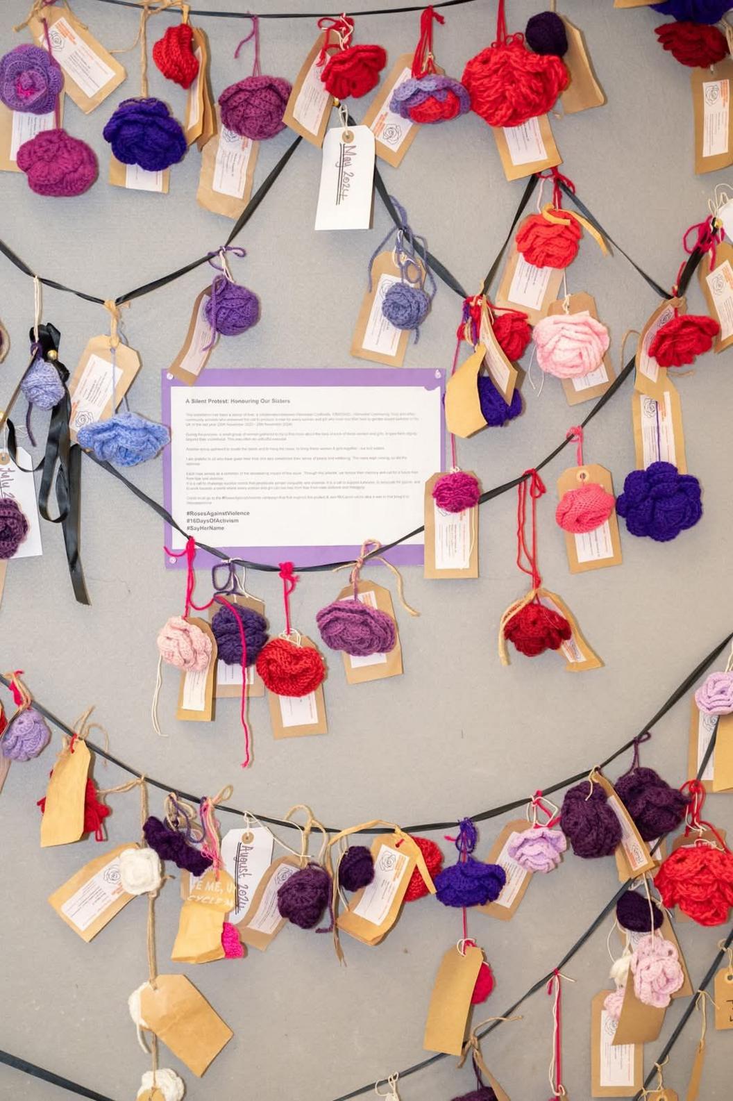 A number of hand-crocheted roses hang on black thread against a grey background. Each rose has a tag attached to it.