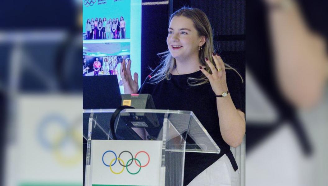 Holly Norman wearing white trousers, a black top, a thin gold watch, dark pink lipstick and dark red nail varnish. She has long, dark blonde hair which is just past her shoulders. She is standing on stage behind a glass podium with the Olympic rings symbol on the front. Behind her there is a large powerpoint presentation full of pictures of the group during the programme. She is smiling and speaking animatedly, with her hands in the air.