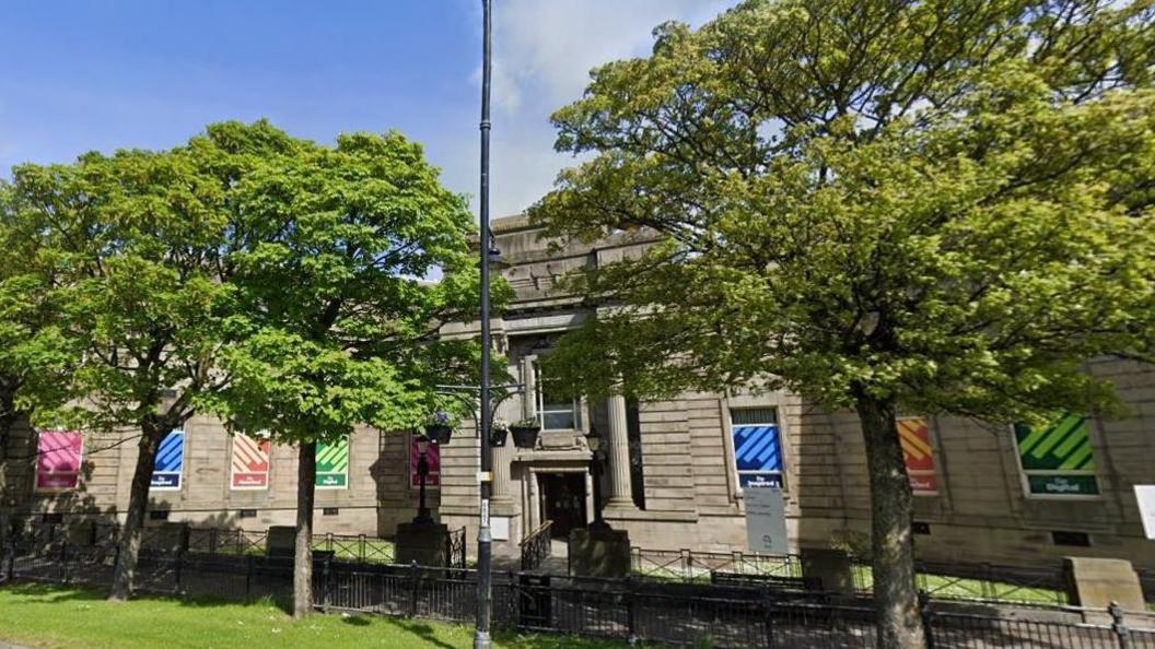 Barrow Library, partially obscured by a row of trees. It is an imposing, though not high, building made from large grey/brown stone. The entrance is flanked by two pillars with a large window above the doors. The windows stretching either side of the central entrance are covered by some sort of promotional materials which are different coloured versions of the same design.