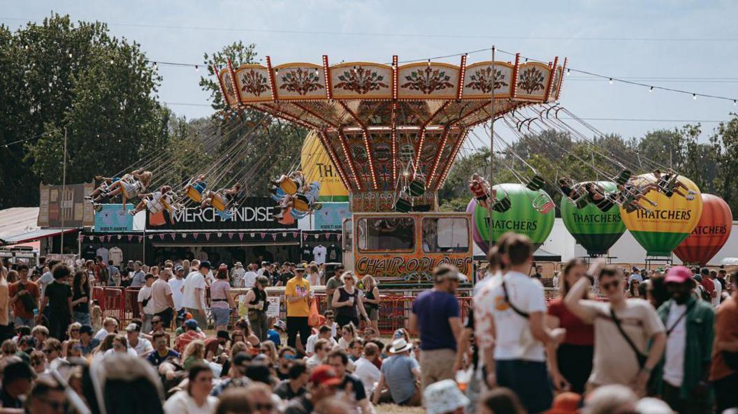 People enjoying attractions at Truck festival. Some can be seen at a merry-go-round.  Thatchers-branded hot balloons branded can be seen in the distance.
