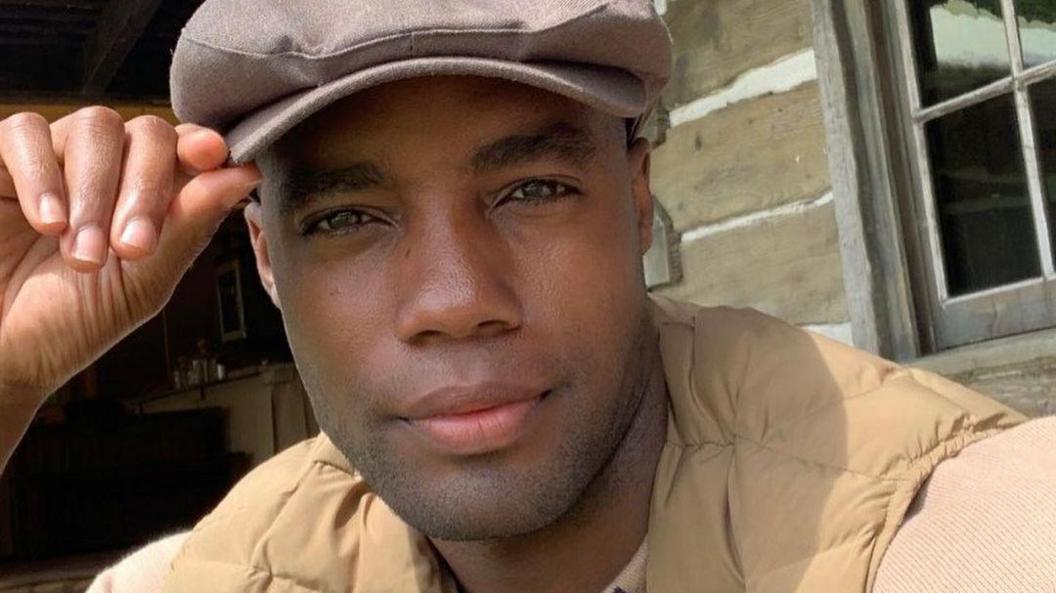 Reece Richards wearing a brown flat cap and beige gilet and ribbed jersey top underneath. He is touching the visor with his hand and is looking directly at the camera.