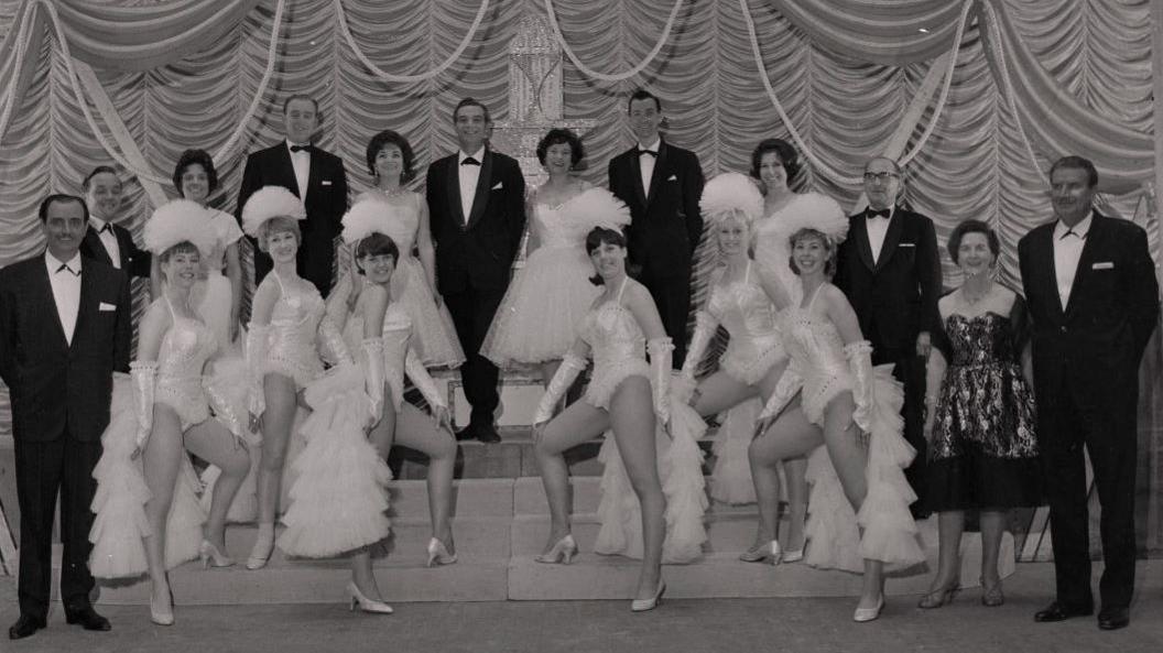 A total of 18 men and women stand on the stage in front of a ruffled curtain. Taken in black and white, the men wear black suits and white shirts and the women wear frilly white dresses, some have head dresses on.