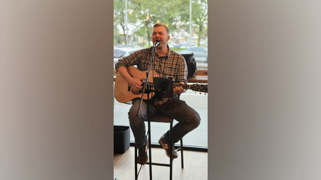 A man sings into a microphone as he plays a guitar. He wears a checked shirt, jeans and trainers. He is sitting on a bar stool in front of a plate glass window, with views over a car park.