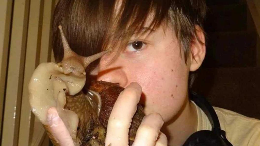 Connor Bouttell holding a large snail, which is bigger than his hand, up to a camera. He is younger than in the other picture of him and has a brown fringe and headphones around his neck. He is wearing a white T-shirt.