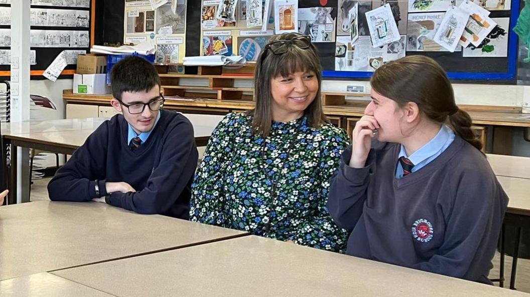 Two pupils sit with a teacher at a High School. They are in a classroom and work is displayed on the wall behind them.