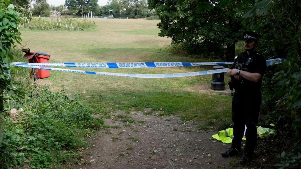 A park in Ipswich cordoned off with police tape. A police officer is also standing nearby.