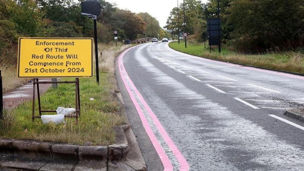 A yellow sign on the grass next to a road informing motorists enforcement measures will commence from 21 October 2024. There are double red lines running along the side of the road.