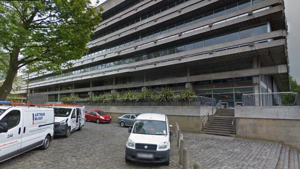 The library is a grey building on several floors. There are a few vans in the foreground.