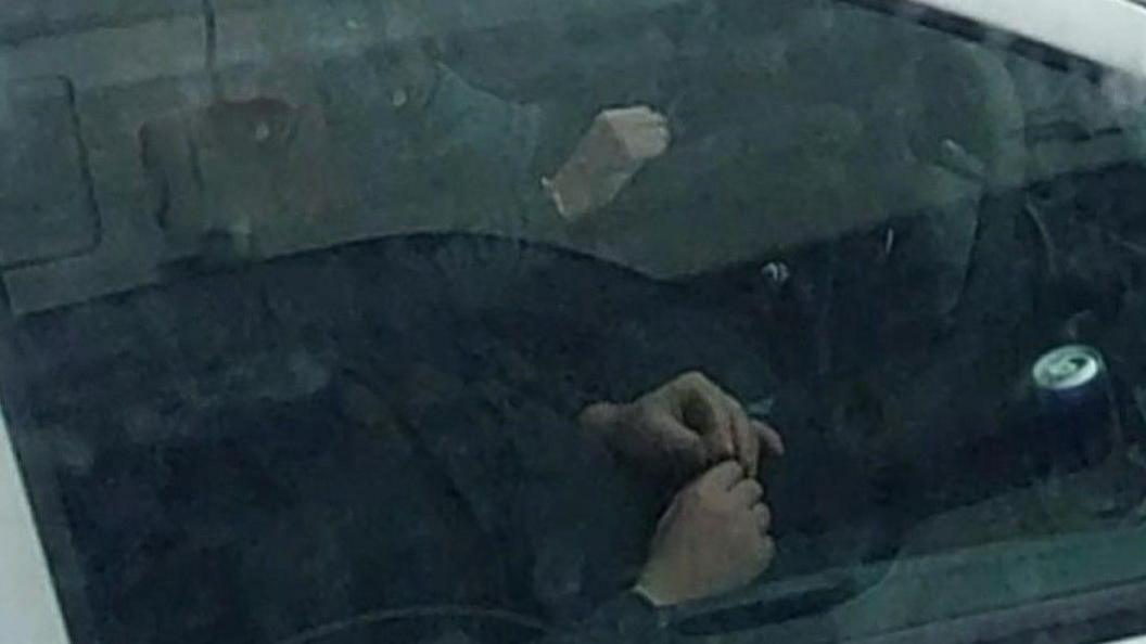 A photo looking into a vehicle, with two hands near the bottom of the image near a steering wheel. Another hand is near the top of the photo.
