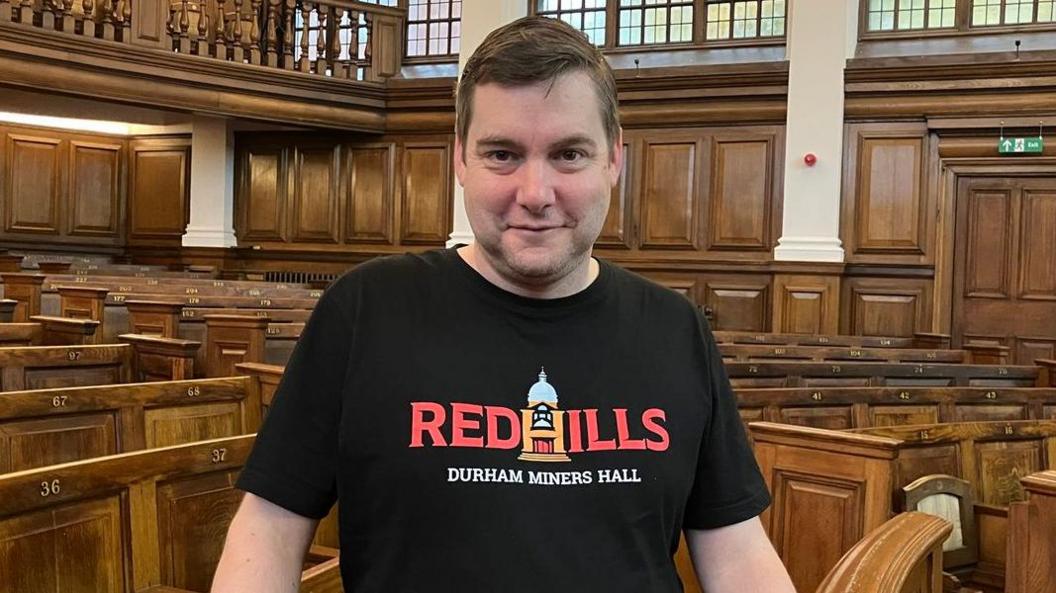 Rob Guest smiling into the camera. He has short, brown hair and is wearing a black t-shirt with the Redhills logo in red letters. He is standing in a large room which is filled with brown wooden chairs. Brown wood lines the walls and there is a balcony.