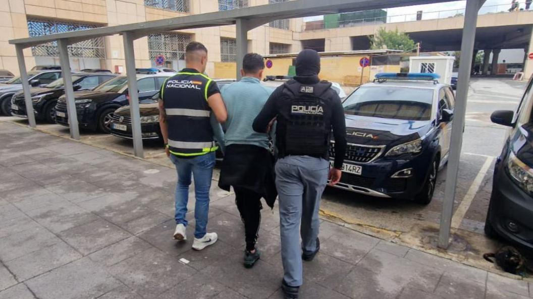 A man in a sky blue long sleeved tshirt is flanked by two police officers wearing bulletproof vests as they walk to a row of police cars.