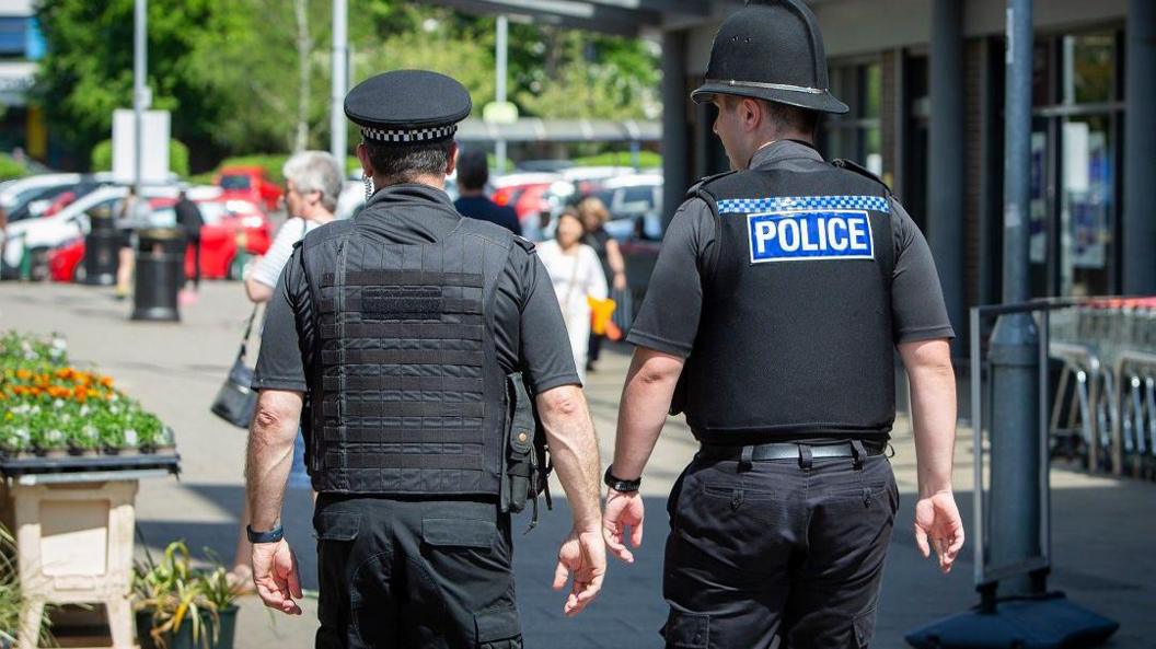 Two police officers in Shirley walking away from the camera, shopping trolley's lined up against a building are to the right.