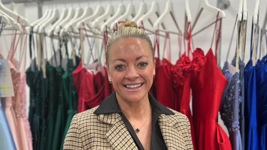 A smiling woman - Kristy Chamberlain - looking straight at the camera. She is wearing a patterned blazer over a black dress and is standing in front of a rail of prom dresses.