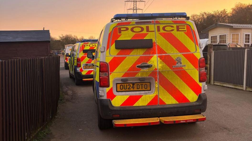 A rear view of three police vans lined up, one behind the other, on a concrete driveway, with a mobile home to the right