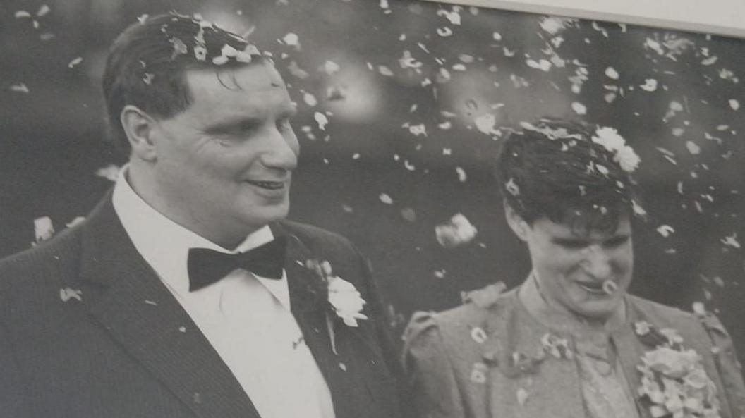 Man with dark suit, white shirt, bow tie and flower on his pocket and with short hair and woman with light jacket and light top with short hair. There is confetti in the air as they both smile on their wedding day.