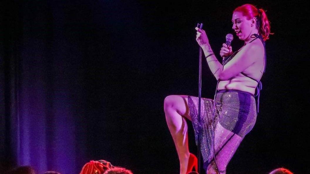 A young woman with red hair scraped back into a ponytail dressed in a sparkly bra and shorts holding a microphone and using its stand to balance on stage. 