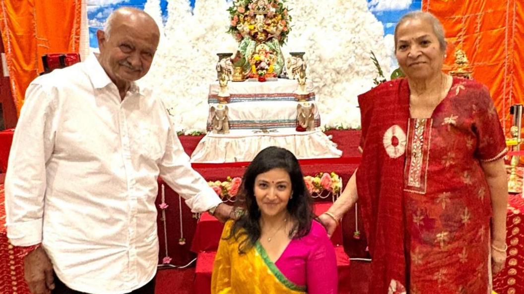 Ekta is sitting down in front of the deities wearing a yellow saree and a pink blouse, with her father in a white shirt (to the left) and mother (right) in a red Indian dress.
