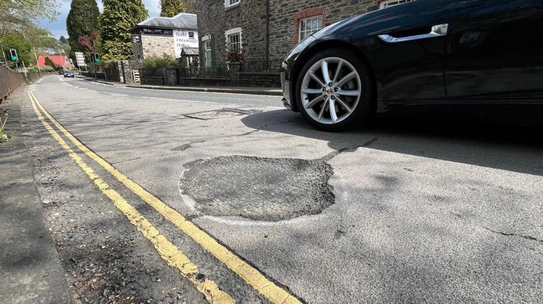 A pothole on a road in Tavistock which has been circled with white paint. There are double yellow lines and a navy blue car is driving past.