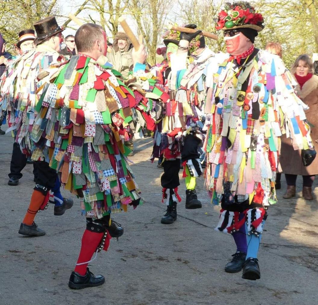 Dancers wear multi-coloured outfits made from pieces of patterned cloth. A crowd of spectators is visible in the background.