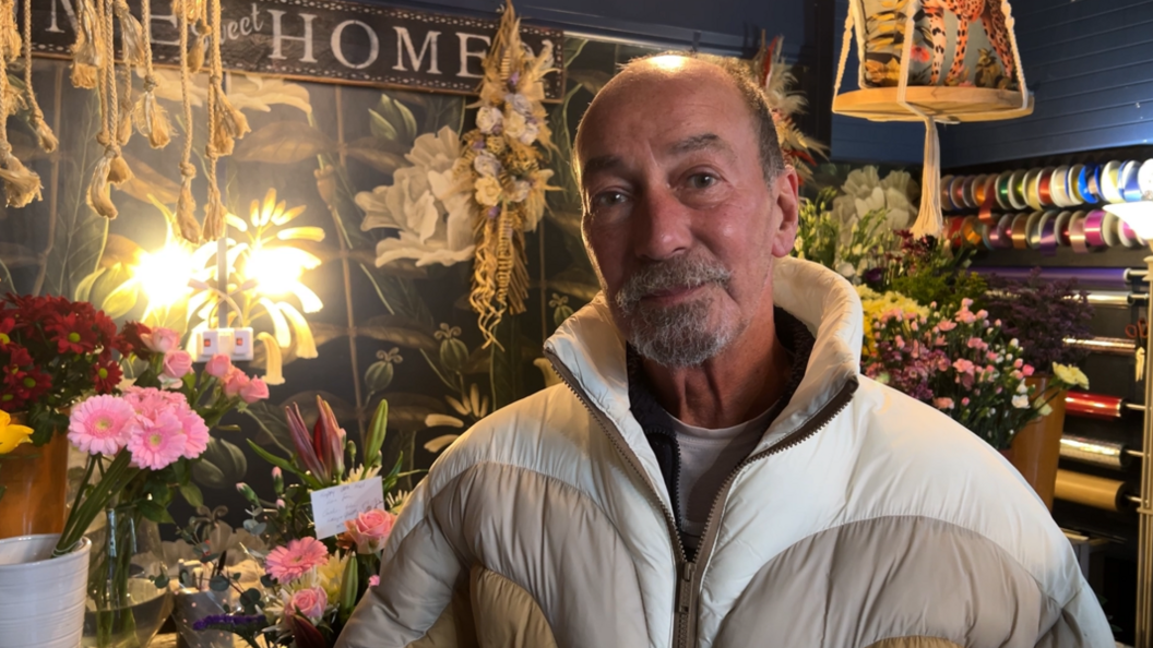 Florist Richard Sharman stands in front of his shop counter, which is full of floral displays, including in pots, baskets and vases. Behind, a feature wall has a painting of flowers.