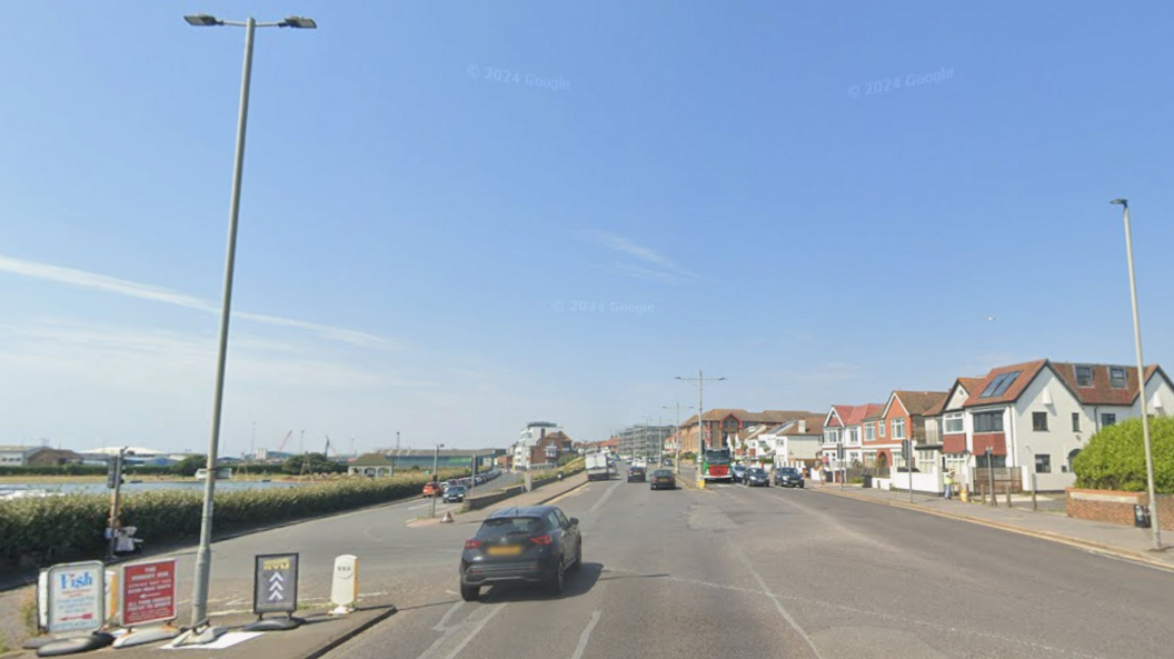 A stretch of road on a sunny day. It is wide and lined by houses. 