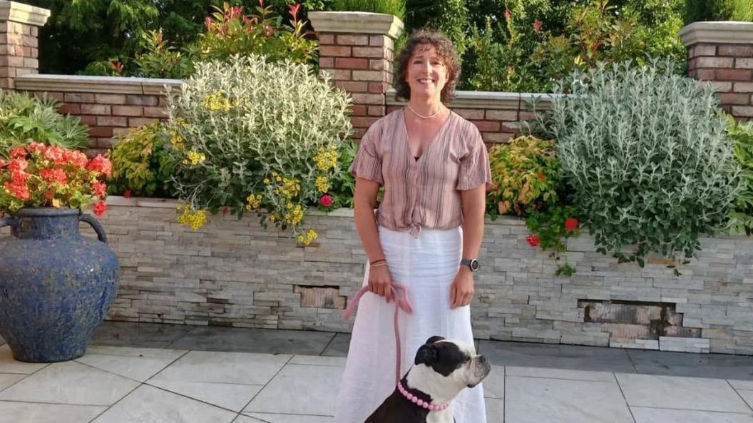 Nicola Joyce stands in a garden wearing a white skirt and pink shirt and holds her bulldog, who has a pink collar that looks like a beaded necklace. She has curly dark hair. In the background is a wall with flowers. 