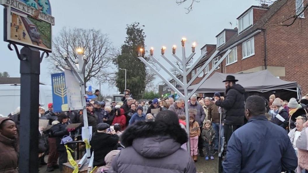 People gathering round a menorah as it is lit by a rabbi. The vandalised menorah is next to it.