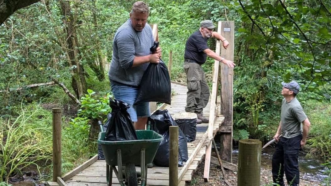 Three men are working in the woods. Two stand on a raised wooden walkway, one of which is lifting a full black bin bag into a wheelbarrow. The other is talking to a man standing below him on the bank of a small stream.