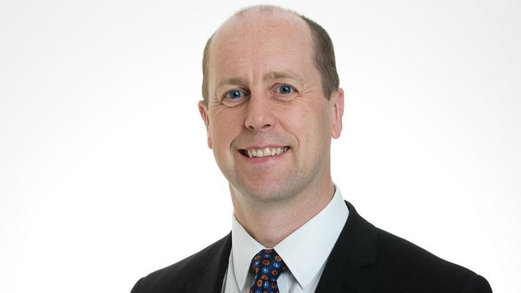 Ryan Scatterty looking directly at the camera, wearing a black suit, white shirt and patterened tie. There is a white background.